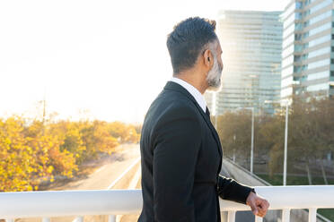 Side view of a mature businessman in a suit standing on a bridge with a pensive look, overlooking an urban city background during daytime. - ADSF54169