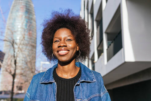 A radiant African American woman with a confident smile stands in an urban landscape, the city's architecture framing her natural beauty - ADSF54173