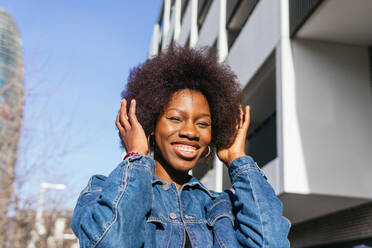 An exuberant African American woman touches her hair and smiles widely, basking in the sunlight against a modern city backdrop - ADSF54174