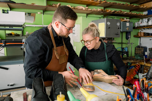 Zwei Schuhmacher, ein Mann und eine Frau, arbeiten gemeinsam an einem Schuh in einer gut ausgestatteten Werkstatt in Österreich, umgeben von Werkzeugen und Maschinen. - ADSF54205