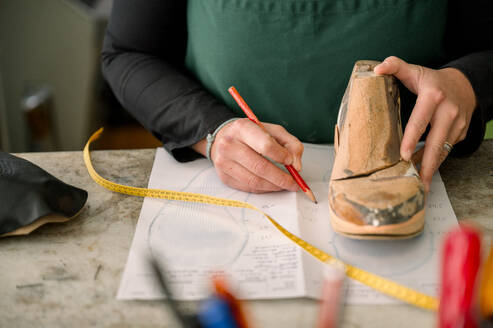 Ein akribisch gekleideter, nicht wiederzuerkennender Schuhmacher in Österreich arbeitet an einem Schuhleisten, misst und markiert mit Präzision für maßgeschneiderte Schuhe. - ADSF54206