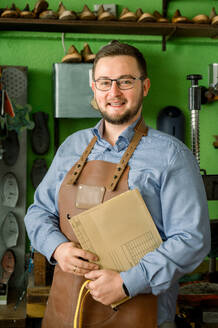 Ein österreichischer Schuhmacher steht stolz lächelnd in seiner Werkstatt, bekleidet mit einem blauen Hemd und einer Lederschürze, in der Hand Schuhmuster. - ADSF54209