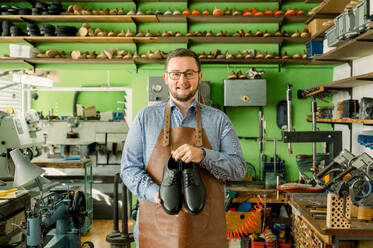Ein fröhlicher Schuhmacher steht in einer gut ausgestatteten Werkstatt in Österreich und präsentiert ein Paar gefertigte Lederschuhe vor einem Hintergrund aus bunten Schuhleisten. - ADSF54210