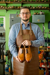 Ein lächelnder Schuhmacher in einer Lederschürze hält ein Paar handgefertigte Schuhe in seiner Werkstatt hoch. - ADSF54213