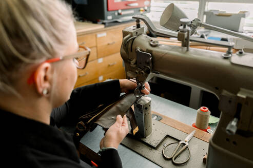 A cropped unrecognizable shoemaker meticulously stitches leather using a sewing machine in a well-equipped workshop in Austria, showcasing craftsmanship. - ADSF54218