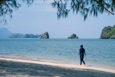 An unrecognizable Muslim woman wearing a burkini is walking along a serene beachfront with tropical islands in the distance. - ADSF54223