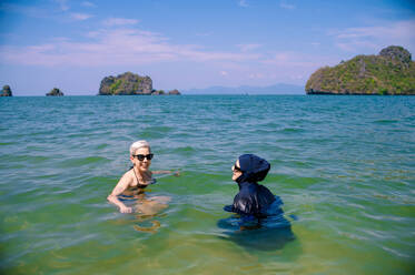 A woman in a burkini alongside a friend in a traditional bikini playfully enjoy the clear waters with a scenic island backdrop. - ADSF54226