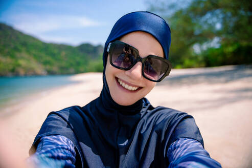 Eine fröhliche Frau, die einen Burkini trägt, macht ein Selfie an einem sonnigen Strand und zeigt vor einer Kulisse aus Sand und Hügeln, dass sie bescheidene Bademode trägt. - ADSF54227