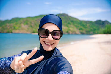 Eine lächelnde Frau in einem Burkini mit Sonnenbrille gibt ein Friedenszeichen und genießt die sonnige Strandkulisse. - ADSF54228