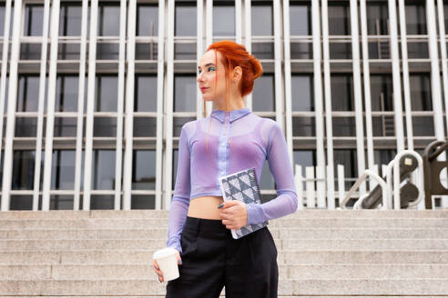 Moderne Geschäftsfrau in lilafarbener durchsichtiger Bluse und schwarzer Hose mit leuchtend rotem Haar, das ihre kühne, unkonventionelle Geschäftskleidung ergänzt, während sie eine Kaffeetasse und ein stilvolles Notizbuch hält - ADSF54233