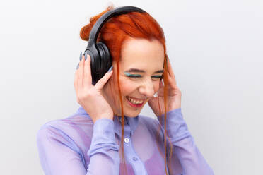 Eine lebhafte, junge kaukasische Geschäftsfrau, die lächelt, während sie mit Kopfhörern Musik hört. Ihre Augen sind sanft geschlossen, mit türkisfarbenem Lidschatten akzentuiert, und ihr Lächeln verrät einen sorglosen Moment - ADSF54234