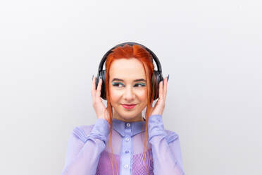 Vor einem weißen Hintergrund steht eine Geschäftsfrau mit leuchtend rotem Haar und einer professionellen lilafarbenen Bluse, die drahtlose Kopfhörer benutzt und wegschaut - ADSF54235