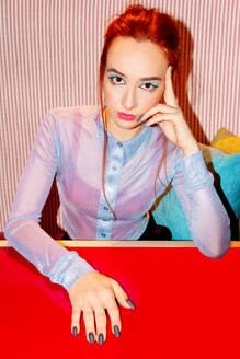 Eine kaukasische Geschäftsfrau mit auffallend rotem Haar blickt nachdenklich in die Kamera. Sie sitzt an einem leuchtend roten Schreibtisch, der mit ihrer lilafarbenen Bluse vor dem Hintergrund einer gestreiften rosa Tapete kontrastiert - ADSF54241