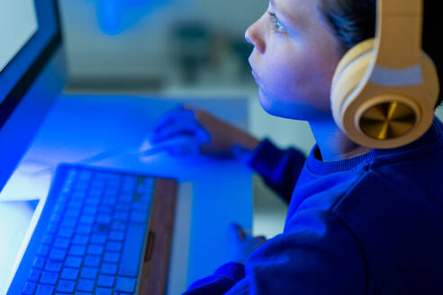 A child is deeply focused on the glowing computer screen, with headphones on, in a darkened room illuminated by the screen's light - ADSF54276