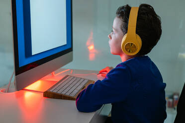 A child is intently using a computer, illuminated by the warm backlight of the monitor, with headphones on - ADSF54277