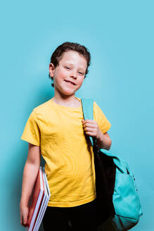 Ein junger Student in einem gelben T-Shirt grinst, während er Lehrbücher in der Hand hält und einen Rucksack trägt, vor einem hellblauen Hintergrund - ADSF54280