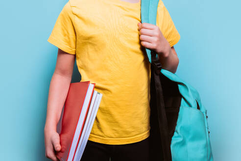 Anonymer junger Student mit gelbem T-Shirt, der Schulbücher in der Hand hält und einen Rucksack trägt, vor einem hellblauen Hintergrund - ADSF54282