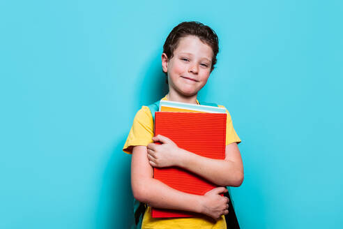 Ein lächelnder Schuljunge in einem gelben Hemd umarmt fest seine Schulbücher, mit einem Rucksack über einer Schulter, vor einem blauen Hintergrund - ADSF54284