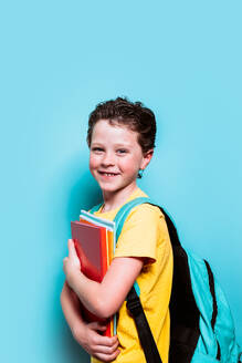 Ein Junge mit strahlenden Augen und einem aufrichtigen Lächeln trägt einen Stapel Bücher und seinen Rucksack und steht vor einem verspielten blauen Hintergrund - ADSF54285