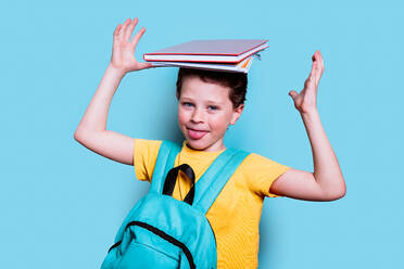 Ein Schuljunge mit frechem Grinsen balanciert Bücher auf seinem Kopf und streckt seine Zunge in einem spielerischen Moment vor einem blauen Hintergrund heraus - ADSF54287