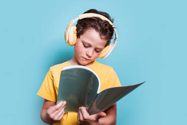 Ein konzentrierter Junge in einem gelben Hemd liest aufmerksam, während er Kopfhörer trägt, vor einem sanften blauen Hintergrund - ADSF54289