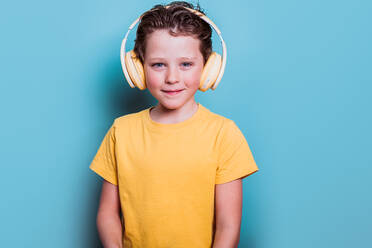 Ein fröhlicher Schuljunge mit einem gelben T-Shirt und Kopfhörern steht vor einem blauen Hintergrund und lächelt freundlich - ADSF54290