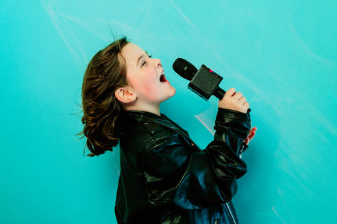 Ein junges Mädchen in einer schwarzen Lederjacke singt ein Lied in ein Mikrofon, das von einem leuchtenden türkisfarbenen Hintergrund angestrahlt wird - ADSF54293