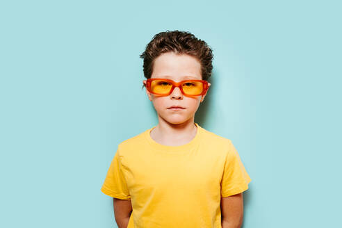 A portrait of a confident young boy wearing vibrant orange sunglasses and a yellow t-shirt, posing against a solid blue backdrop - ADSF54300