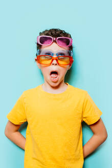 Ein verspielter Junge trägt mehrere Sonnenbrillen vor einem leuchtenden türkisfarbenen Hintergrund und macht einen überraschten Gesichtsausdruck - ADSF54301