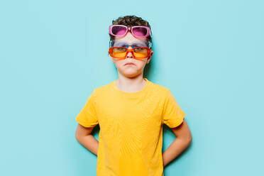 Ein Junge in einem gelben Hemd trägt selbstbewusst drei Sonnenbrillen auf einem hellblauen Hintergrund, der eine spielerische Haltung zeigt - ADSF54302