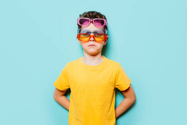 Young boy in a yellow shirt confidently sports three pairs of sunglasses on a bright blue background, showcasing a playful attitude - ADSF54302