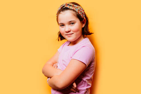 Ein junges Mädchen in einem lässigen T-Shirt und einem bunten Stirnband posiert selbstbewusst mit verschränkten Armen vor einem leuchtend gelben Hintergrund - ADSF54305