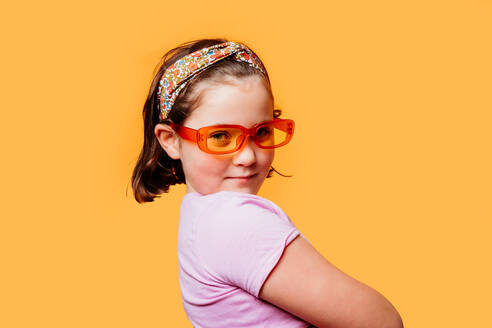 Nettes junges Mädchen mit orangefarbener Sonnenbrille und einem bunten Stirnband posiert selbstbewusst vor einem orangefarbenen Hintergrund - ADSF54306