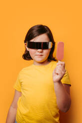 A child in a yellow shirt and futuristic sunglasses holding an ice cream on a yellow background - ADSF54307