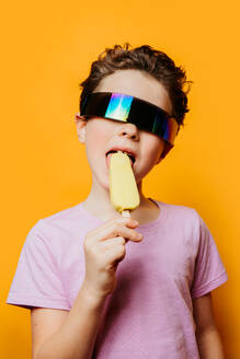 Child with funky sunglasses enjoys an ice cream against a vibrant orange background - ADSF54308