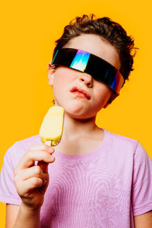 Ein Kind mit stylischer, übergroßer, futuristischer Sonnenbrille probiert ein Eis vor einem warmen Hintergrund und drückt damit eine coole Sommerstimmung aus - ADSF54309