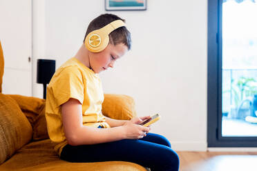 Ein fokussierter Junge sitzt auf einem Sofa, trägt gelbe Kopfhörer und benutzt ein Smartphone, mit einem gemütlichen Hausinterieur im Hintergrund - ADSF54316