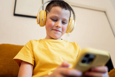 Junge im gelben Hemd genießt Musik über seine Kopfhörer, während er ein Smartphone hält - ADSF54317