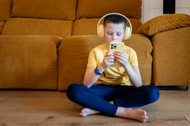 A focused young boy sits cross-legged on a wooden floor, wearing headphones and holding a smartphone, with a sofa in the background - ADSF54320