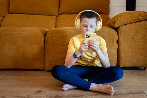 Ein konzentrierter Junge sitzt im Schneidersitz auf einem Holzboden, trägt Kopfhörer und hält ein Smartphone in der Hand, im Hintergrund steht ein Sofa - ADSF54320