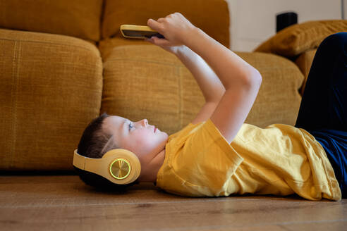 Kleiner Junge, der zu Hause auf dem Boden liegt, Kopfhörer benutzt und ein Smartphone in der Hand hält - ADSF54321