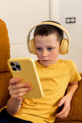 Kleiner Junge im gelben T-Shirt, der zu Hause mit Kopfhörern in sein Smartphone vertieft ist - ADSF54322