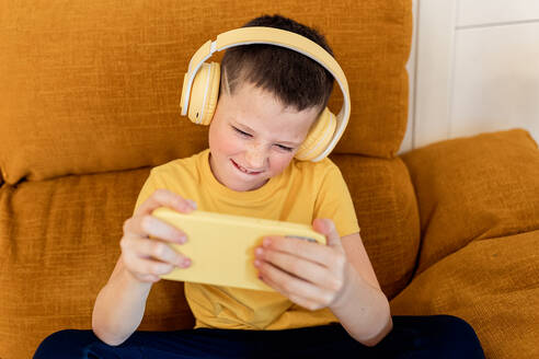 Ein Junge in einem gelben T-Shirt spielt lächelnd ein Spiel auf seinem Smartphone und sitzt mit Kopfhörern auf einer orangefarbenen Couch - ADSF54323
