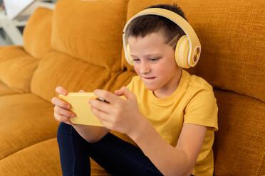 Ein konzentrierter Junge mit gelben Kopfhörern spielt auf einem Sofa sitzend Spiele auf einem Smartphone - ADSF54324