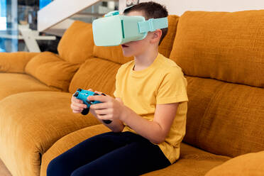Ein Junge sitzt auf einem Sofa und spielt ein VR-Spiel, er trägt ein Headset und hält einen Controller - ADSF54325