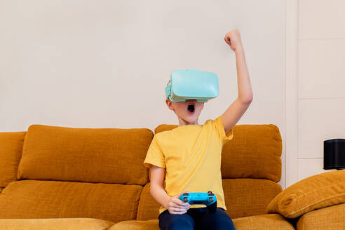 Ein kleiner Junge zelebriert eine Siegesgeste, während er mit einem VR-Headset spielt, während er zu Hause auf einer Couch sitzt - ADSF54326