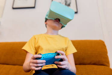 Ein junger Mensch sitzt auf einem Sofa und spielt mit einem VR-Headset und einem Game-Controller, völlig eingetaucht in ein virtuelles Spiel - ADSF54327