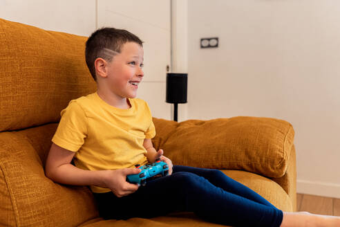 Ein kleiner Junge sitzt bequem auf einer warm getönten Couch und spielt vergnügt mit einem Videospiel-Controller. Er zeigt ein Lächeln, das von echter Freude und Engagement für seine Freizeitbeschäftigung zeugt - ADSF54330