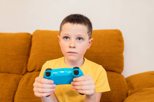 Ein konzentrierter Junge spielt Videospiele mit einem blauen Controller in der Hand, auf einer braunen Couch sitzend - ADSF54331