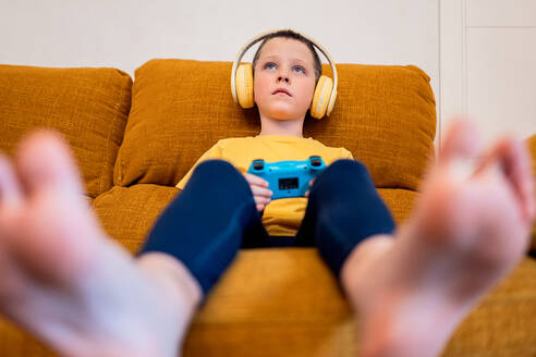 Ein konzentrierter Junge sitzt auf einer braunen Couch, trägt gelbe Kopfhörer und hält einen blauen Game-Controller in der Hand, vertieft in ein Videospiel - ADSF54335